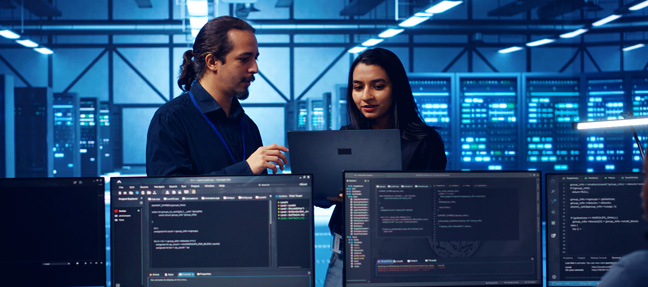 Two NOC Technicians collaborating with laptops and monitors displaying network monitoring dashboards inside a high-tech server room.